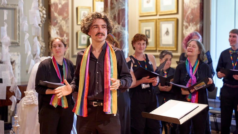 A group of people in black clothing with rainbow coloured accents singing inside a highly decorative interior with gold framed pictures and marbled walls